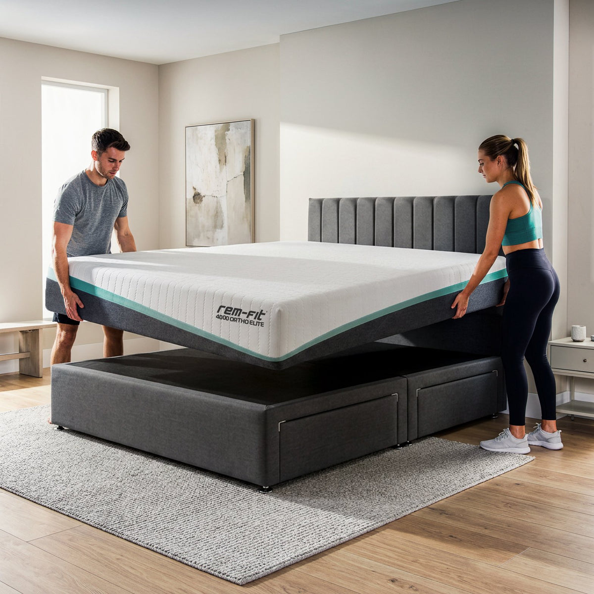 A couple relaxes in bed on a gray upholstered platform bed with white bedding and a visible mattress. The room is bright and modern with a white rug, bedside lamp, wall art, and a plant.